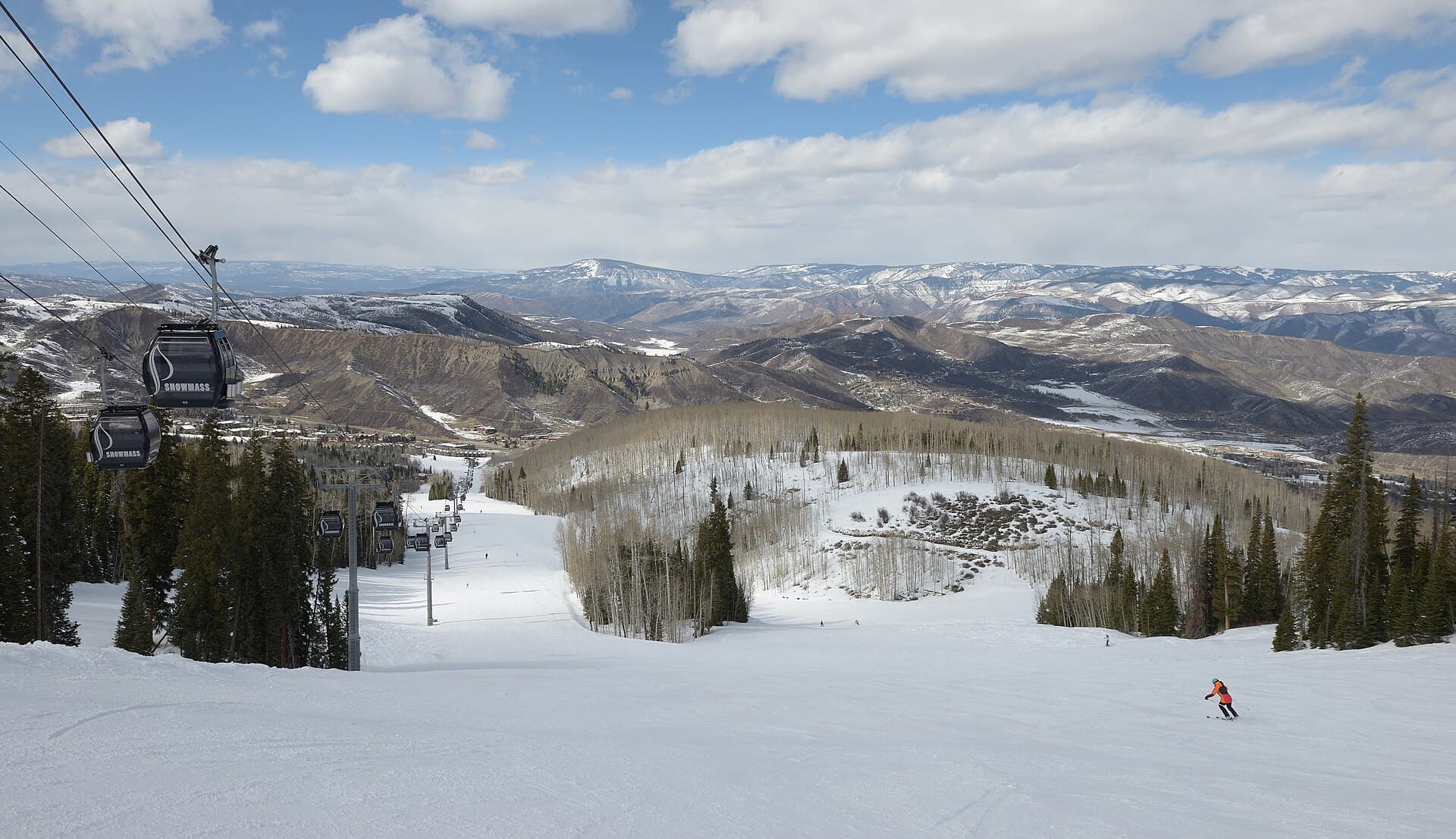 A skier descends beneath the Snowmass gondola with Colorado mountains beyond